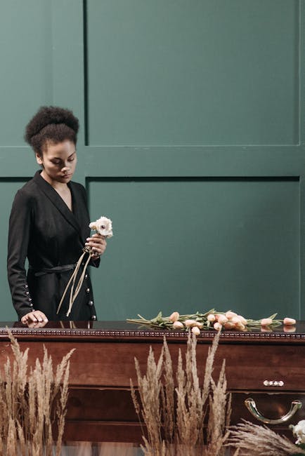 A young woman with curly dark hair styled in an updo, wearing a black blazer with pearl-button details on the sleeves, stands beside a wooden casket, holding a small white flower bouquet with a beige