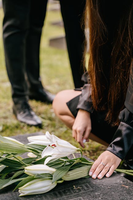 A person dressed in black, kneeling at a graveside with floral arrangements consisting of white lilies and green foliage, their hands gently resting on the ground near the flowers. In the background,