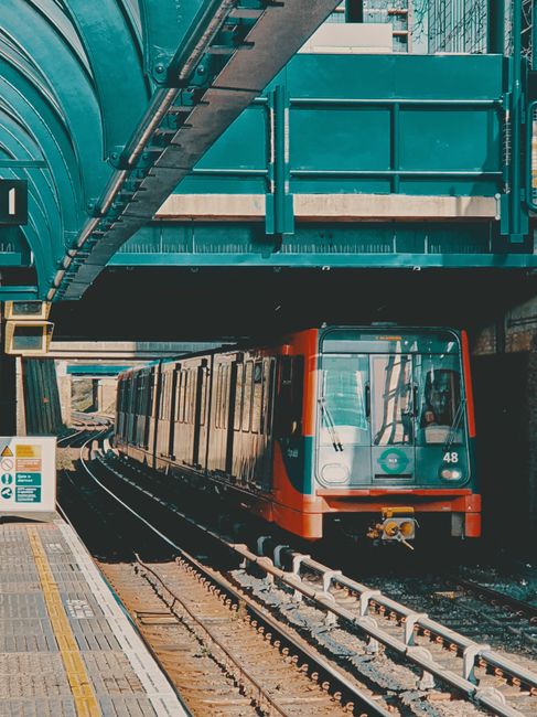 This image depicts a modern train approaching a station platform beneath a teal-colored overhead footbridge with glass paneling. The train has a sleek, streamlined design with a white body accented by