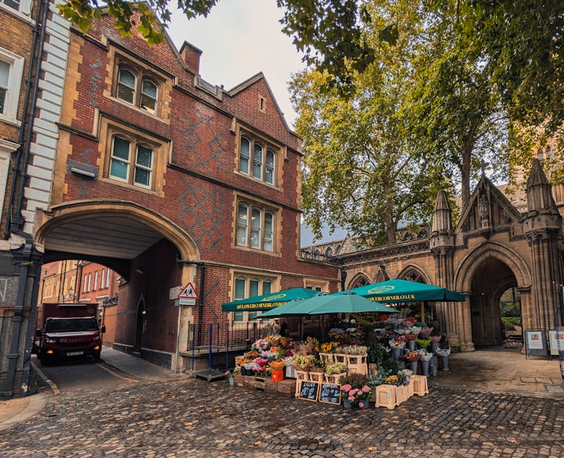 A bustling outdoor flower market situated on a cobbled street near a historic red brick and stone archway, featuring a vibrant display of fresh flowers arranged in wooden crates and buckets under a la