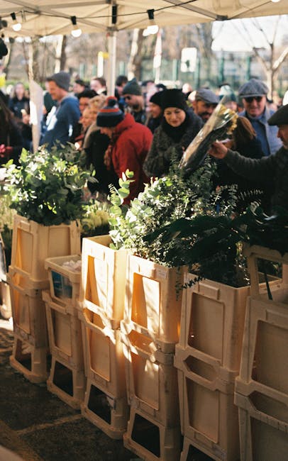 A busy outdoor flower market scene featuring several wooden crates filled with vibrant, fresh floral arrangements and greenery. The flowers include lush green foliage, white blossoms, and hints of oth