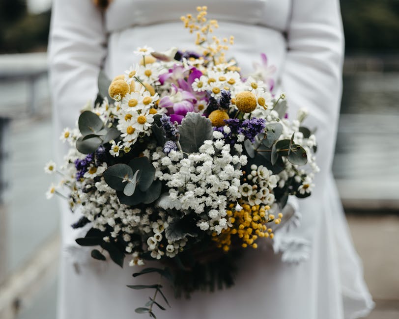 A person dressed in white holds a large floral bouquet featuring a variety of fresh flowers, including white daisies with yellow centers, purple and white lisianthus, small yellow mimosa blooms, and d