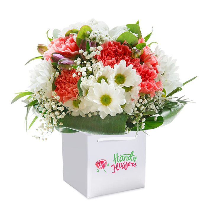 Elegantly nestled in a gleaming white gift bag, this bouquet radiates the vibrant, contemporary style found on the streets of Shadwell. The focal point is a collection of lavish coral, peach, and deep red carnations, their plush, ruffled petals forming a tapestry of rich texture and colour. Stems of glowing white daisies punctuate the design, each with a striking chartreuse-yellow center-echoing the bright, fresh spirit of flower stalls in nearby Watney Market. Interwoven sprays of delicate white gypsophila float amid the main blooms, creating an airy softness that catches the eye and soothes the senses. On the bouquet's left, a few unopened buds shyly peek through, dusted with a hint of purple, adding subtle depth and anticipation. Supporting all is a base of lush, deep green foliage; glossy, broad leaves create a strong foundation and evoke the greenery of local canalside gardens. The white bag is finished with sturdy ribbon handles, ready for gifting or same-day delivery along The Highway or Jamaica Street. Each detail is crisply illuminated against a white background so the intricate forms and living colours take center stage. Here, the vibrancy, care, and professionalism of Shadwell's florists are on vivid display-this bouquet is a celebration piece, suited for birthdays, congratulatory moments, or simply uplifting someone special in the neighbourhood.