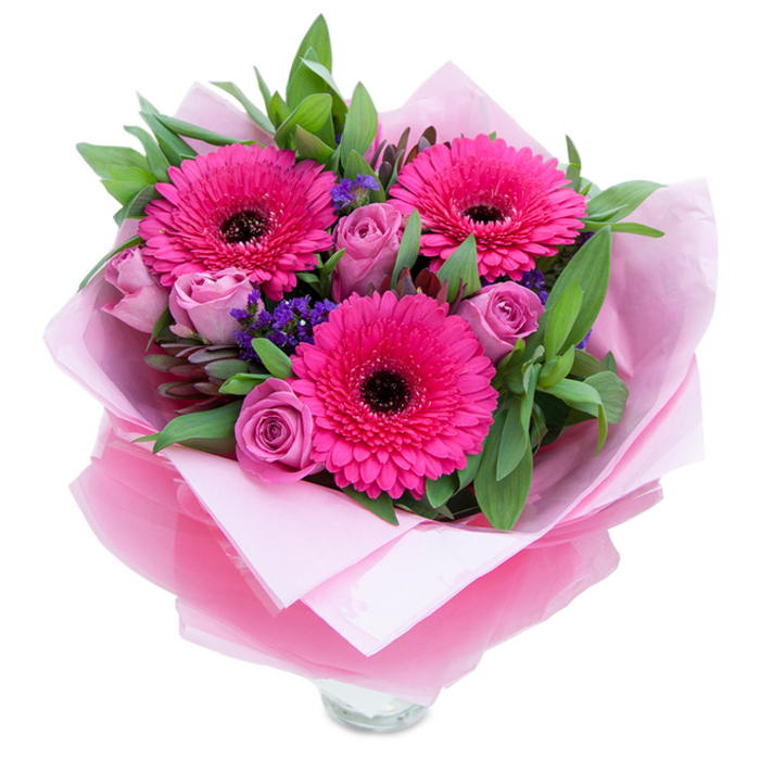 There is a playful elegance to this bouquet that reads like springtime in miniature. Three fuchsia Gerbera daisies command attention with their saturated colour and many-layered petals, each centred by a dark disk that anchors the composition. Around them, roses in a muted lavender-pink or dusty-rose hue sit in a poised, bud-like state - petals curled inward with the soft resilience of new blooms - offering a romantic counterbalance to the Gerberas' boldness. Intermittent sprays of purple statice add texture and a sprinkling of cool colour, their fine clusters giving the eye a place to rest between the larger flowers. A generous arrangement of glossy, broad foliage - suggestive of salal or eucalyptus - builds volume and a fresh, garden-scented base that supports the blooms. The bouquet is wrapped with tactile care: a soft matte pale-pink tissue paper wraps the stems, and over that a layer of slightly crinkled, translucent fuchsia-pink cellophane creates a delicate shimmer that makes the whole presentation feel like a thoughtful gift. The clear glass vase rim peeks from the lower portion, indicating the arrangement is prepared for immediate display on a mantel, bedside table or dining centrepiece. The fragrance is unobtrusive and pleasant - a hint of rose perfume with a clean, green note from the gerberas and foliage. As a florist familiar with Shadwell and the nearby Docklands, I picture this bouquet being delivered to mark an anniversary, to brighten a flat on Cable Street, or to send as a warm 'thinking of you' to a neighbour - a confident, joyful arrangement that balances drama and tenderness.