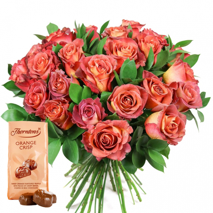 Centered against a pristine white ground, a sumptuous bouquet of roses presents a warm chorus of colour - from concentrated coral and deep peach in the buds' cores to softer salmon, pink and cream on the outer petals - many carrying a subtle ombré or bicolour effect that reads like soft brushwork across each bloom. The arrangement is densely packed so the individual flower heads meld into a single, opulent dome; interspersed foliage of dark green leaves, likely eucalyptus or salal, provides sculpted negative space and a cooling, verdant foil to the rosy tones. Petal textures are velvety and layered, catching the light in gentle highlights that suggest a fresh-cut fragrance lingering just out of frame. Lower-left in the composition, a light beige Thorntons Orange Crisp pouch stands upright with the brand logotype and bold product name visible, and a small pictorial of two glossy truffles reinforcing the pairing; two unwrapped milk chocolate truffles sit before the pouch, their smooth, rounded surfaces reflecting the studio light. The stems appear gathered and trimmed beneath the flower head, hinting at a professional hand-tie prepared for hand delivery. The visual language here - warm colour, tactile petals, and a gourmet chocolate accent - speaks to celebration and comfort, making this an ideal choice for marking birthdays, Valentine's moments, or Mother's Day in Shadwell, near Cable Street or the old Docklands warehouses, communicating care through texture, scent and refined simplicity.